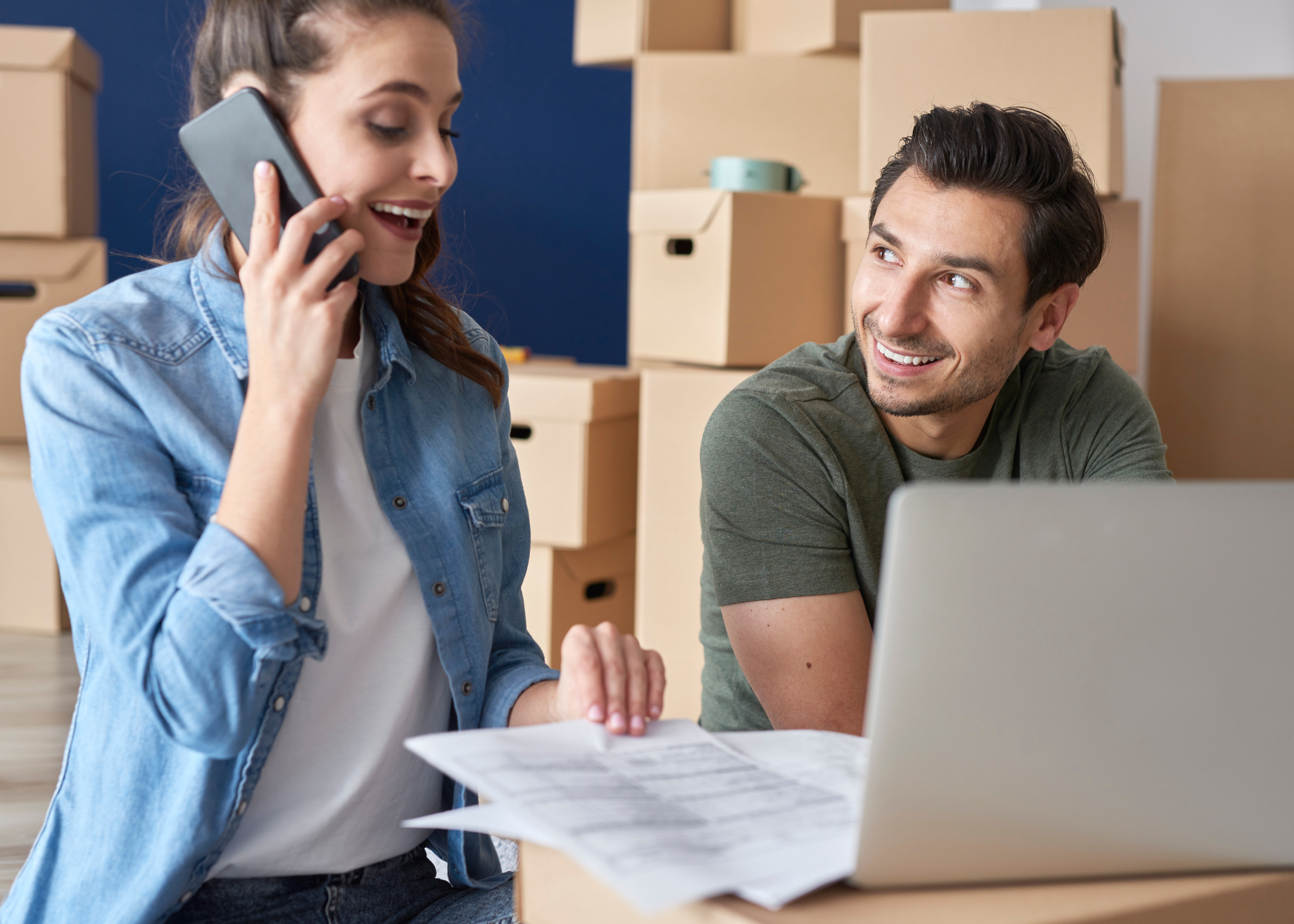 Young couple in home with moving boxes on phone and computer with financial documents
