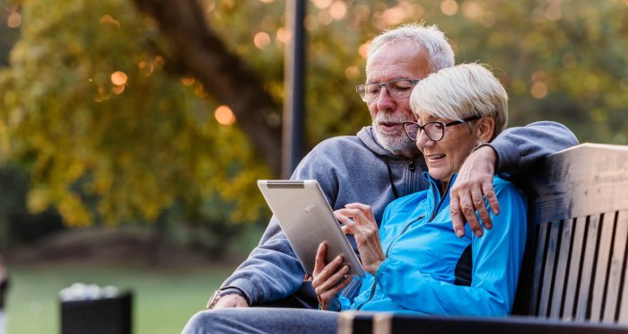 Smiling active senior couple sitting on a park bench looking at tablet computer
