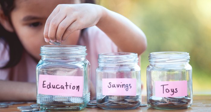 Young girl putting coins into glass bottles labeled education, savings and toys in the garden - saving money for the future