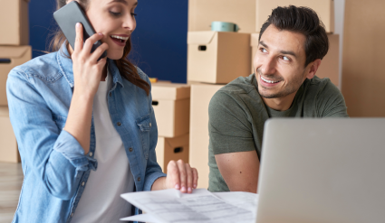 Young couple in home with moving boxes on phone and computer with financial documents