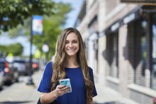 Young woman standing on sidewalk smiling and holding mobile phone
